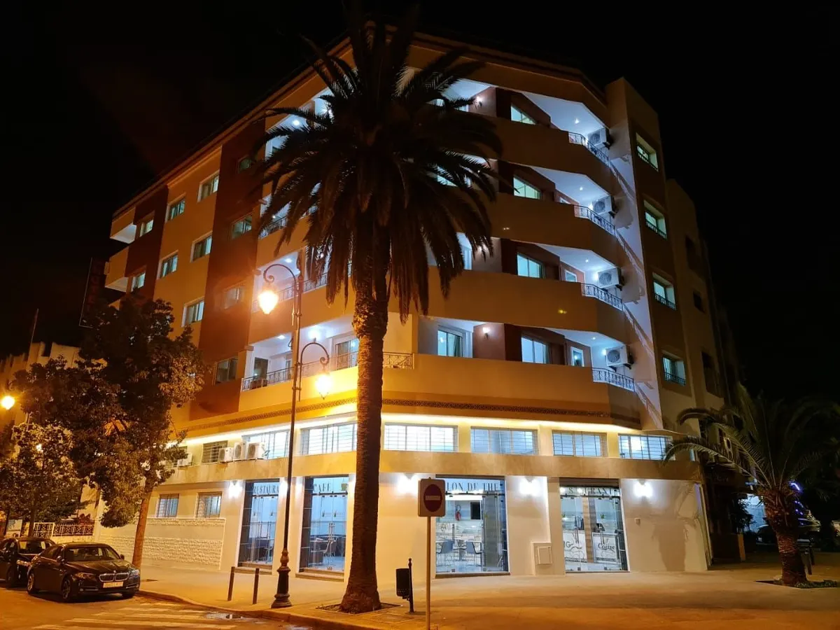 L'angle de l'immeuble la nuit, sous un grand palmier : rez-de-chaussée éclairé, lampadaires, voitures garées