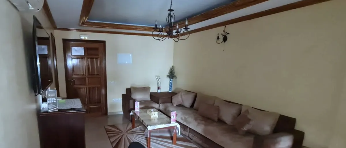 Salon avec canapé en L, table basse sur un tapis rayé, télévision murale et porte d'entrée en bois
