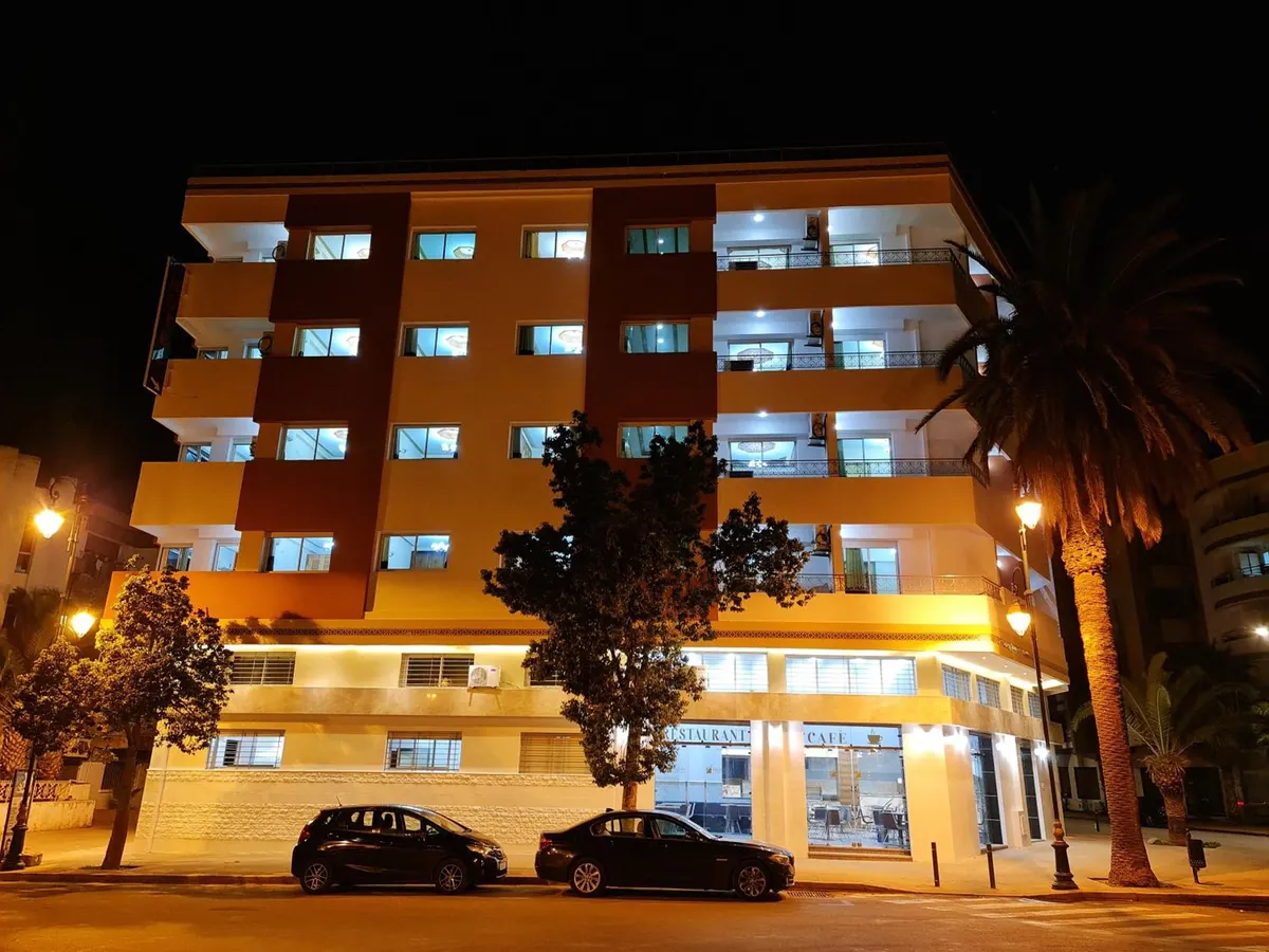 L'immeuble de nuit : cinq étages de balcons éclairés, le café allumé au rez-de-chaussée, un palmier au coin